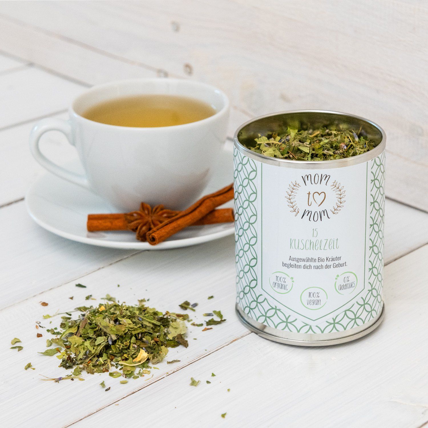 Tasse Tee, Zimtstange, Dose mit Kräutermischung. Aufschrift: mom to mom Kuschelzeit, 100% vegan, Bio.