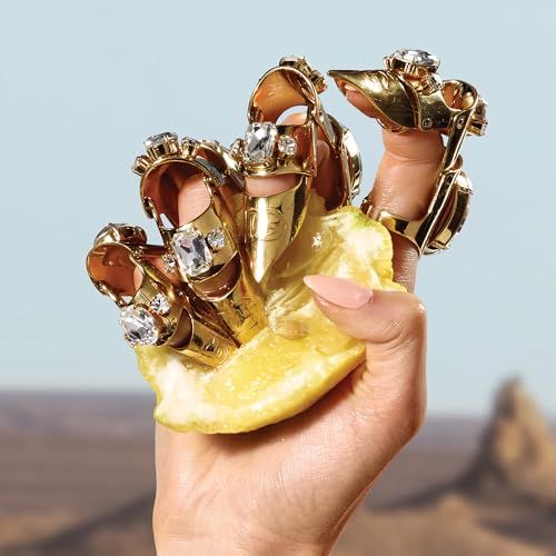 Hand mit goldenen Ringen, besetzt mit Steinen, hält Zitronenscheibe. Hintergrund: Wüste.
