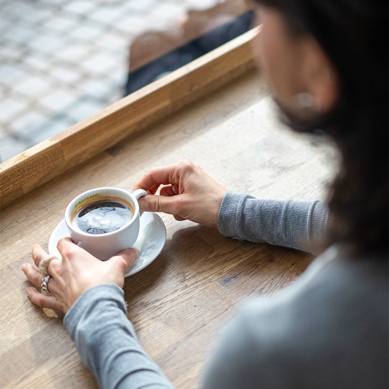 Person hält eine Tasse Kaffee auf einem Unterteller. Holzoberfläche. Fenster im Hintergrund.
