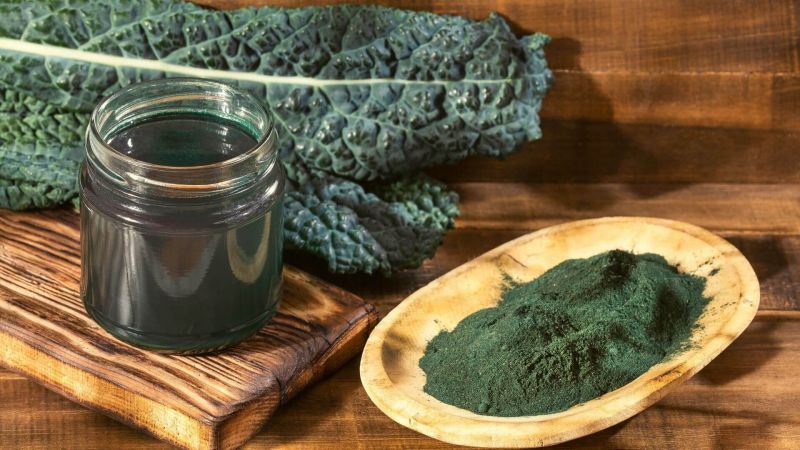 Glas mit Flüssigkeit, daneben ein Holzschälchen mit grünem Pulver. Im Hintergrund Grünkohlblätter.