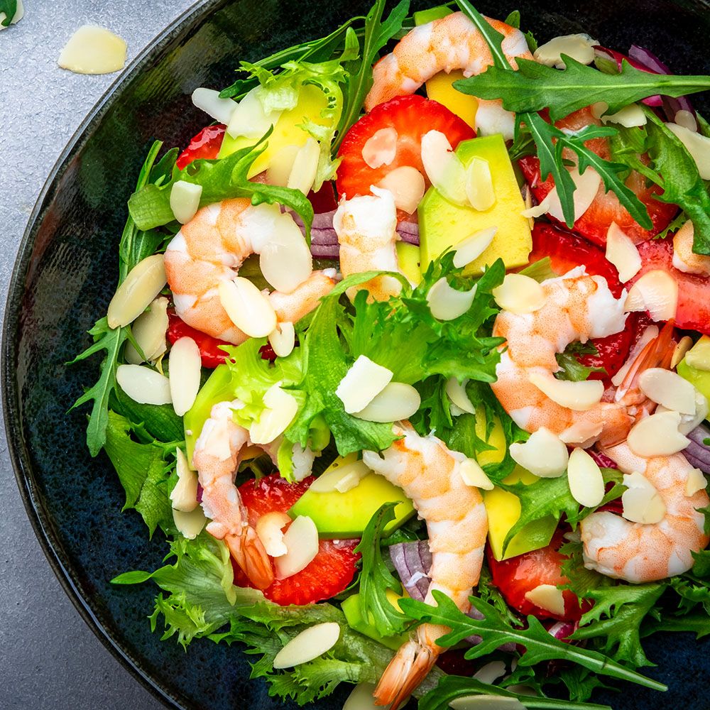 Salat mit Garnelen, Erdbeeren, Avocado und Mandelsplittern in einer schwarzen Schüssel. Auf grauem Untergrund.
