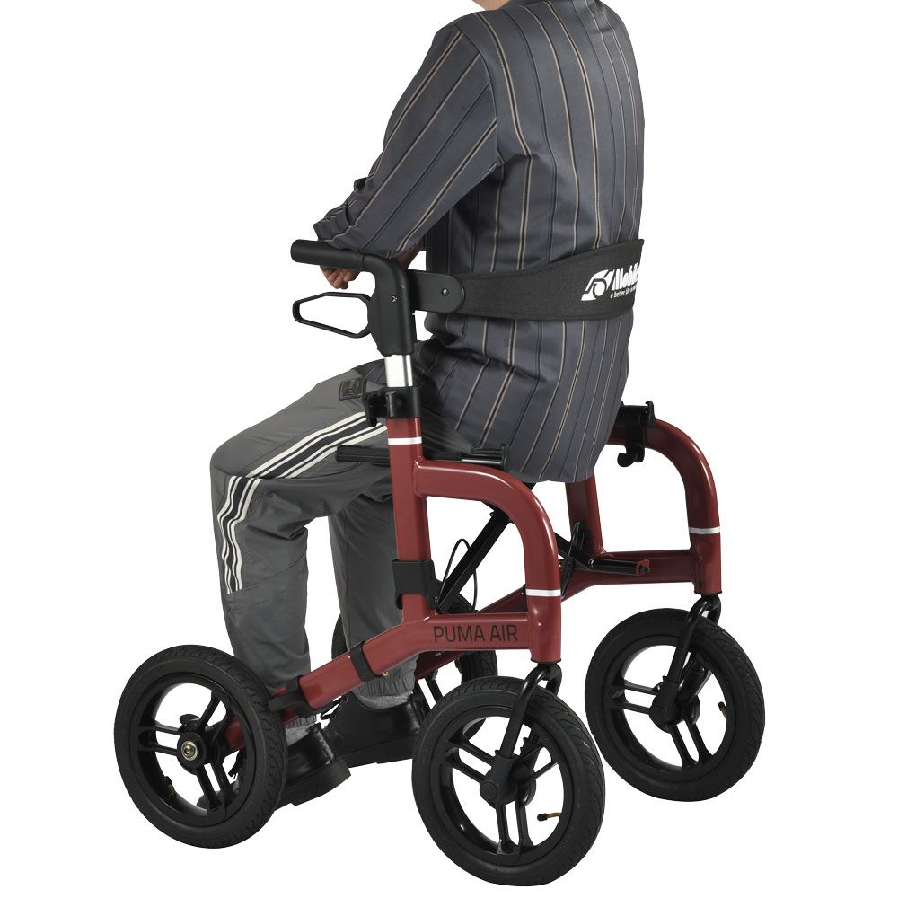 Person sitzt auf Rollator Puma Air. Roter Rahmen, schwarze Räder. Sitzfläche, Rückenlehne. Person trägt Hose und Jacke.