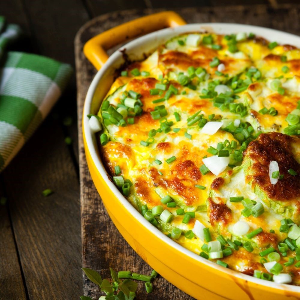 Gelber Auflauf in ovaler Form. Mit grünen Zwiebeln garniert. Auf Holzbrett. Grünes Tuch links.