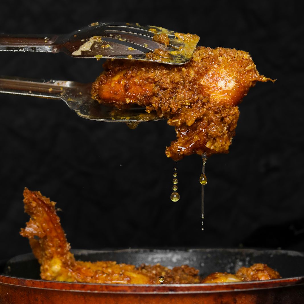 Hähnchenflügel mit goldbrauner Kruste, Zange hält ihn. Öl tropft. Im Topf weitere Flügel.