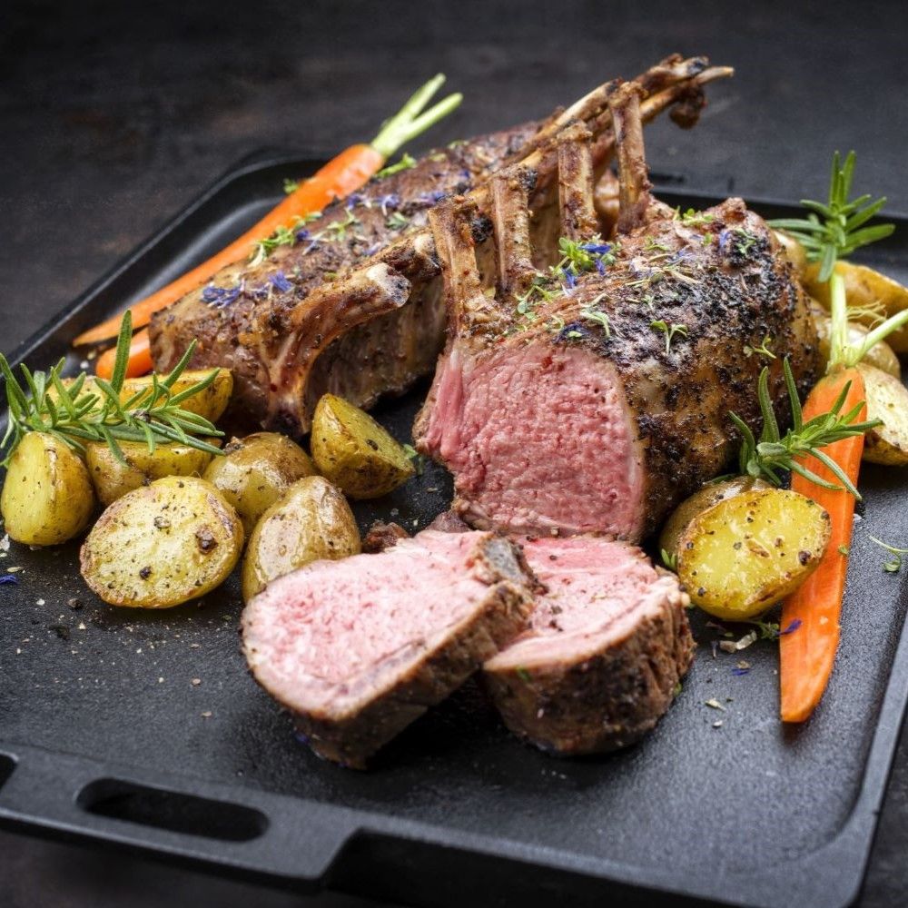 Gebratenes Lammkarree auf schwarzem Tablett, mit Kartoffeln, Karotten und Rosmarin. Fleisch rosa, angeschnitten.