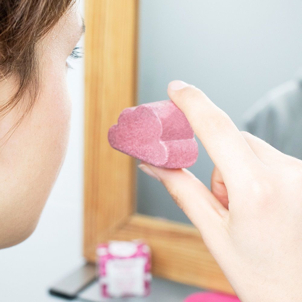 Person hält festes Gesichtsreinigungsmittel in Wolkenform vor Spiegel. Rosa Produkt, Wolkenform.