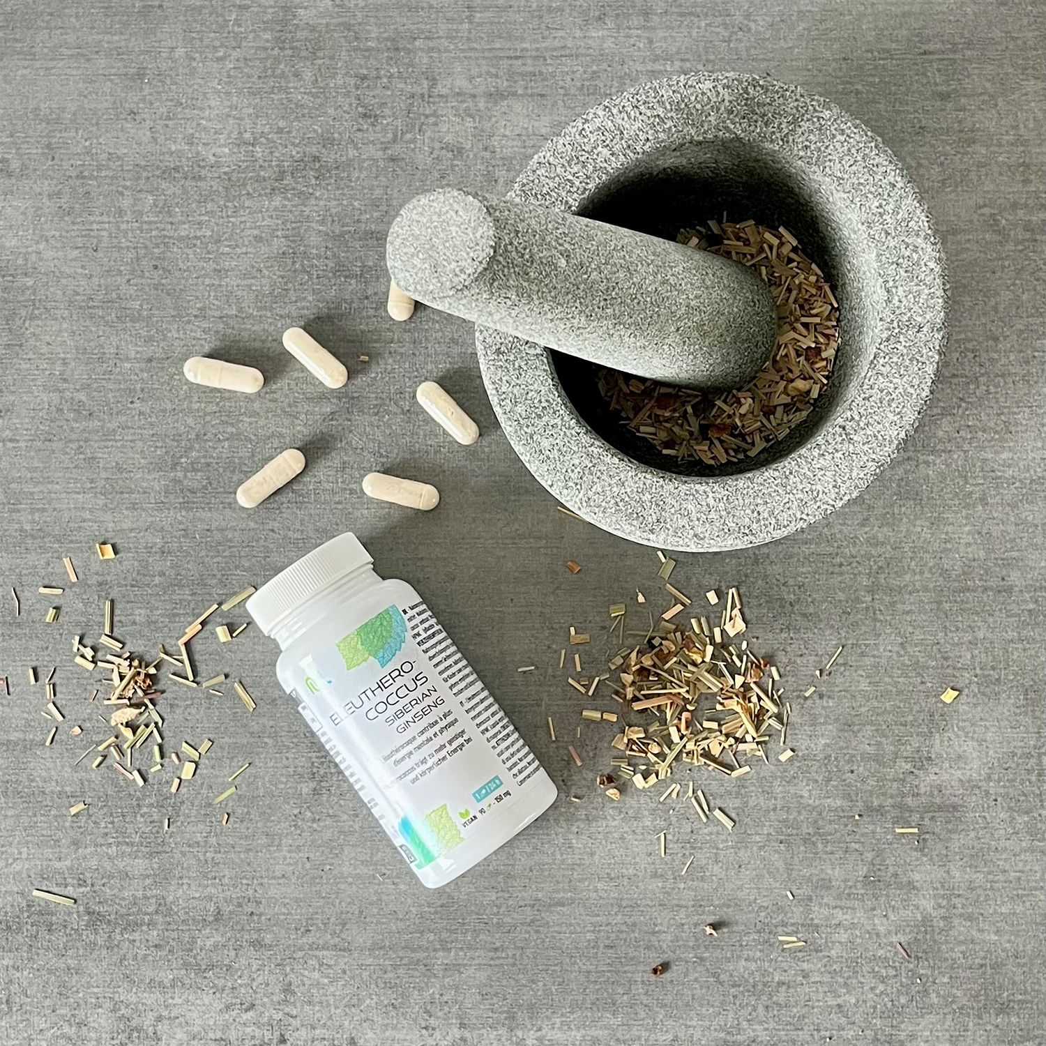 Weiße Flasche mit Kapseln neben Mörser und Kräutern. Aufschrift: Eleutherococcus Siberian Ginseng. Kräuter und Kapseln verstreut.