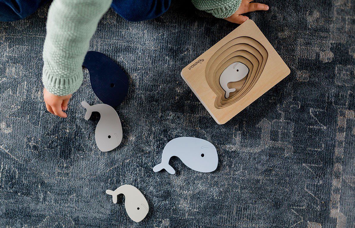 Kindsgut Wal-Puzzle mit Wal-Puzzleteilen. Mehrere Wal-Puzzleteile liegen neben dem Puzzle. Kind greift nach einem Teil.