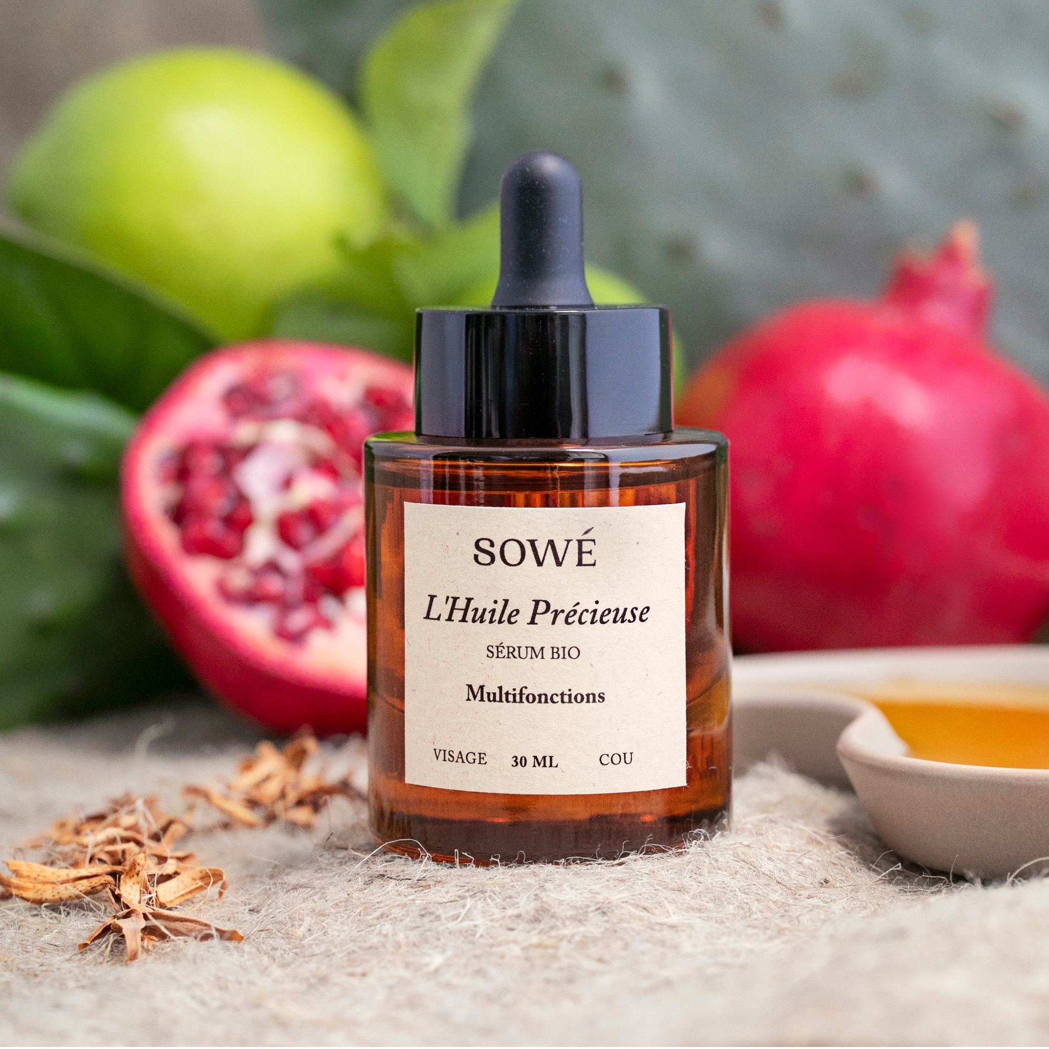 Braune Glasflasche mit Pipette. Etikett mit Produktnamen und Text. Im Hintergrund Granatapfel und Limette.