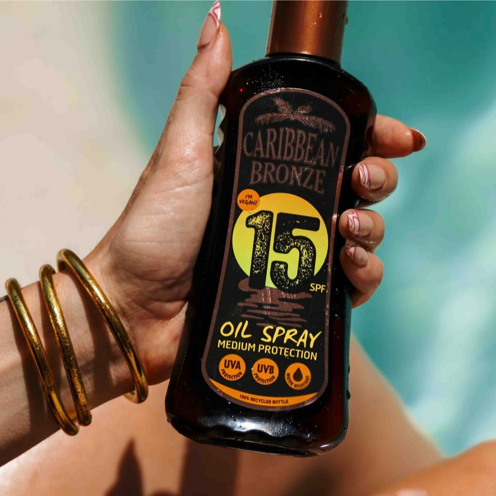 Braune Flasche in Hand gehalten. Aufschrift: Caribbean Bronze, 15 SPF, Oil Spray, Medium Protection. Goldene Armreifen.