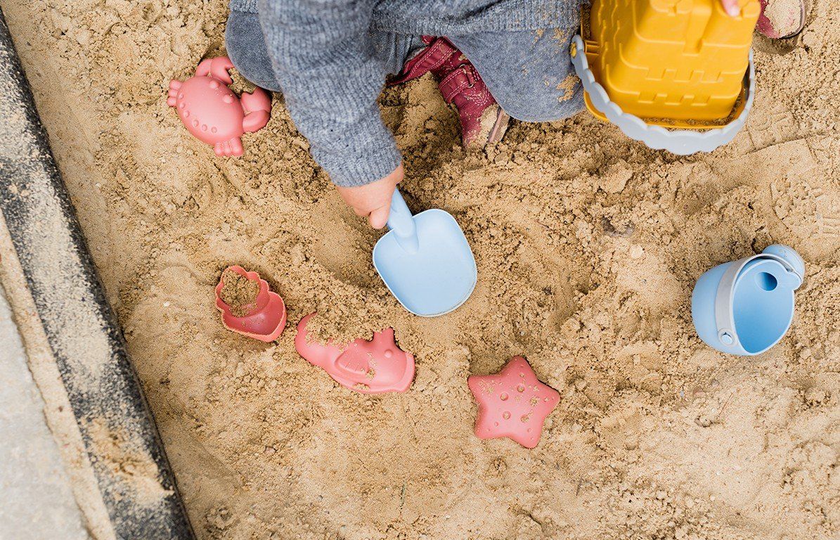 Kind spielt im Sandkasten mit gelbem Eimer, blauer Schaufel und rosa Formen. Sandspielzeug auf Sand.