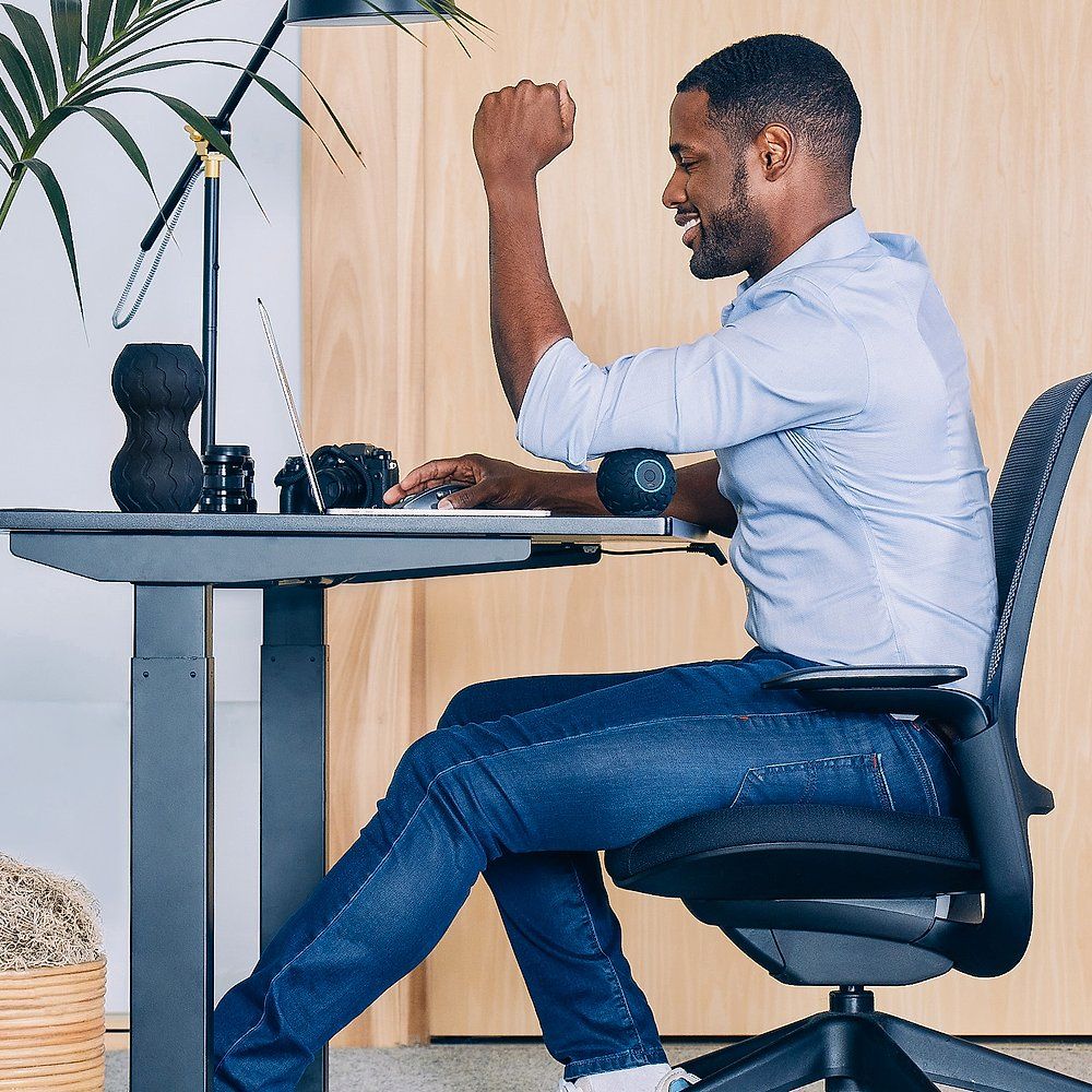 Mann sitzt an einem Schreibtisch, der Massageball am Arm. Er hebt die Faust. Laptop und Lampe sind sichtbar.
