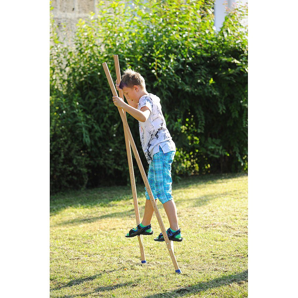 Ein Junge steht auf Stelzen im Gras. Die Stelzen sind aus Holz und haben blaue Gummifüße. Der Junge trägt Schuhe.