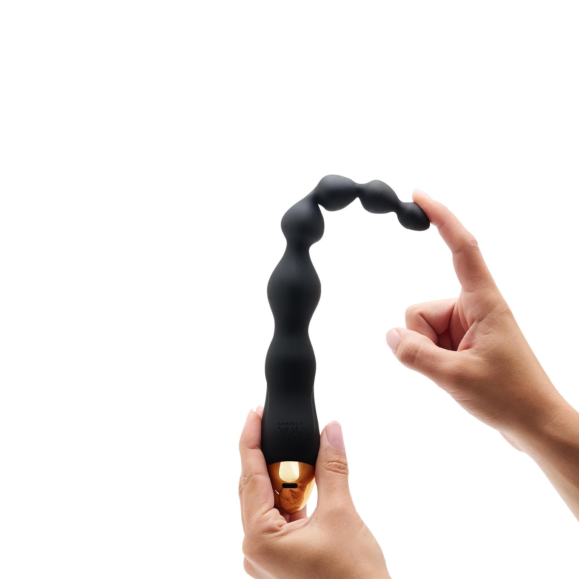 Schwarze, vibr. Anal-Kette, teilweise gebogen, wird von Händen gehalten. Goldfarbenes Ende sichtbar.