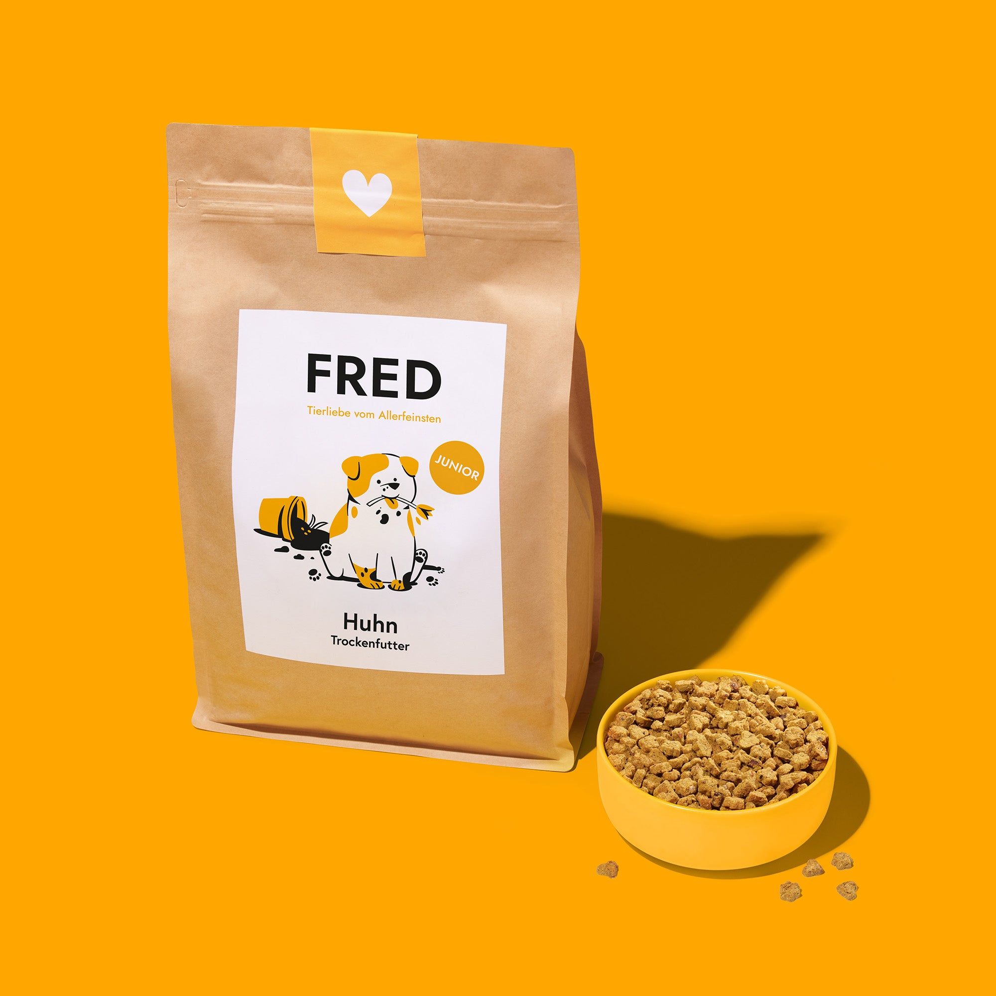 Braune Papiertüte mit Aufdruck und gelbe Schüssel mit Trockenfutter. Logo FRED, Text: Huhn Trockenfutter, Junior. Illustration: Hund.