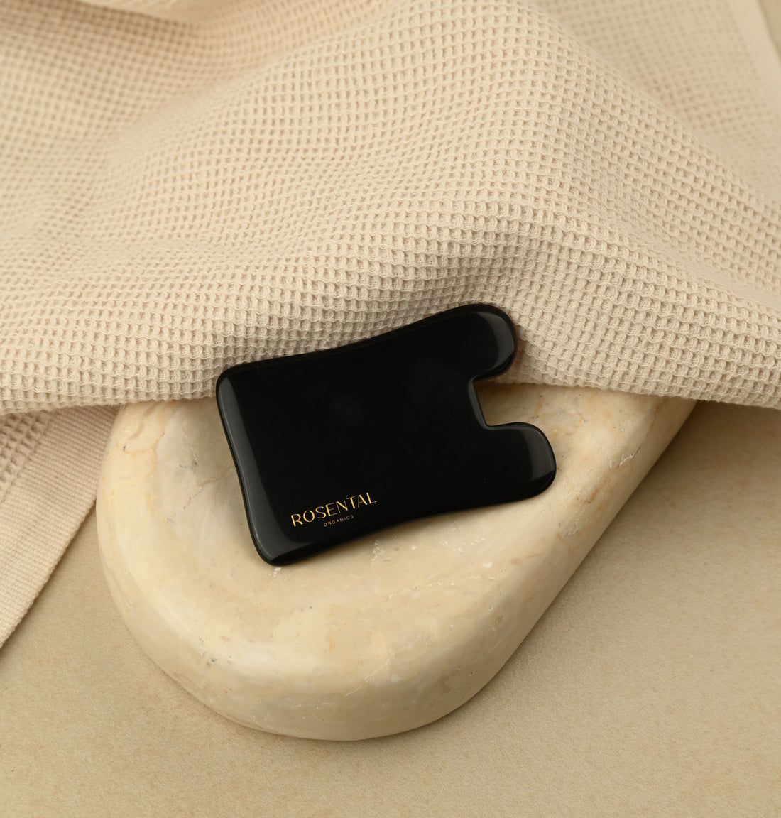 Schwarzes Gua Sha auf einem Stein. Liegt auf einem beigen Handtuch. Goldener Schriftzug: ROSENTAL ORGANICS.