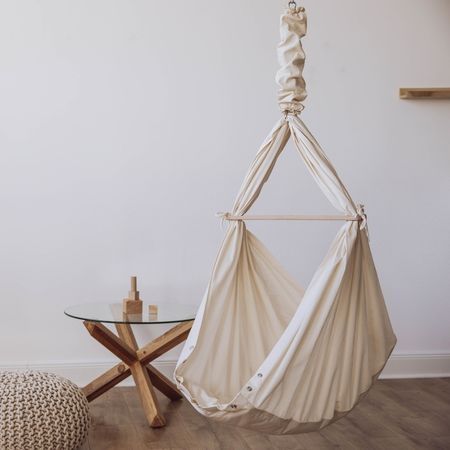 Babyhängematte in beige. Hängematte hängt an Feder. Steht neben Tisch und Hocker.