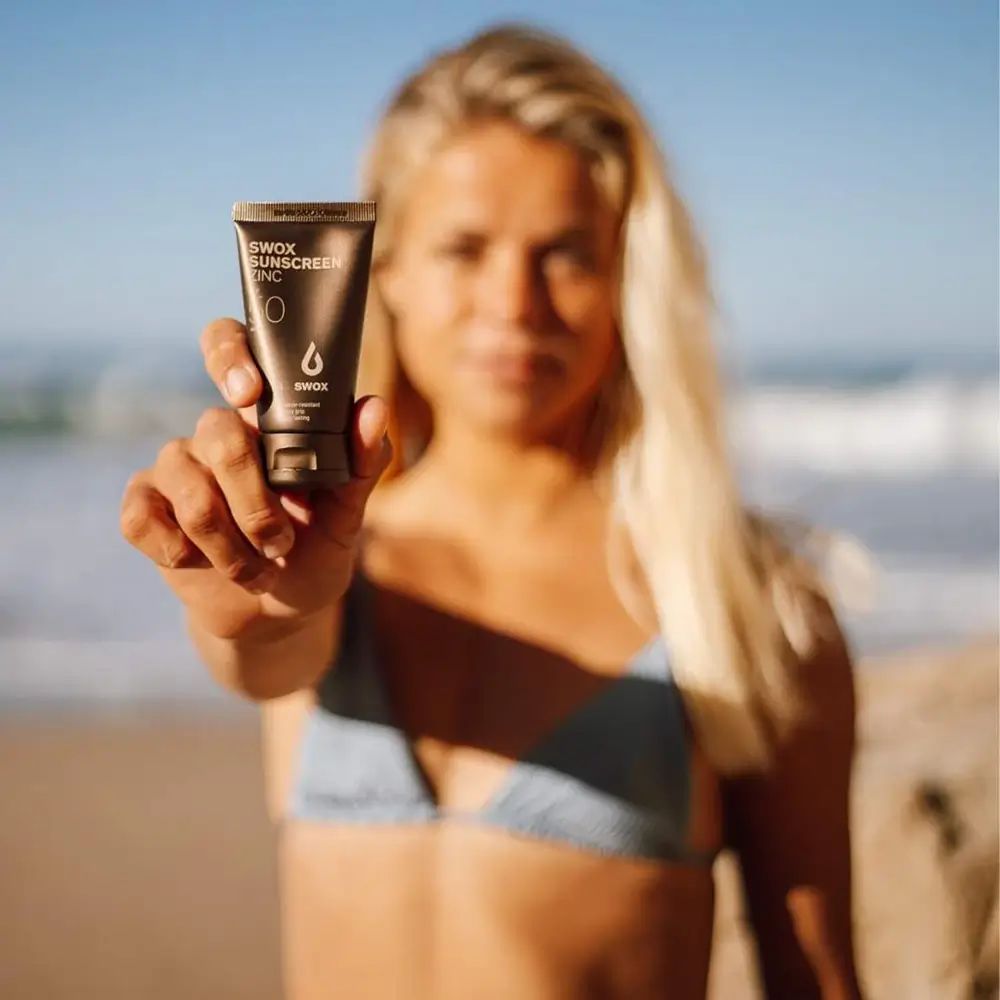 Person hält eine braune Tube SWOX SUNSCREEN ZINC SPF50. Frau mit blonden Haaren am Strand. Logo und Text sichtbar.