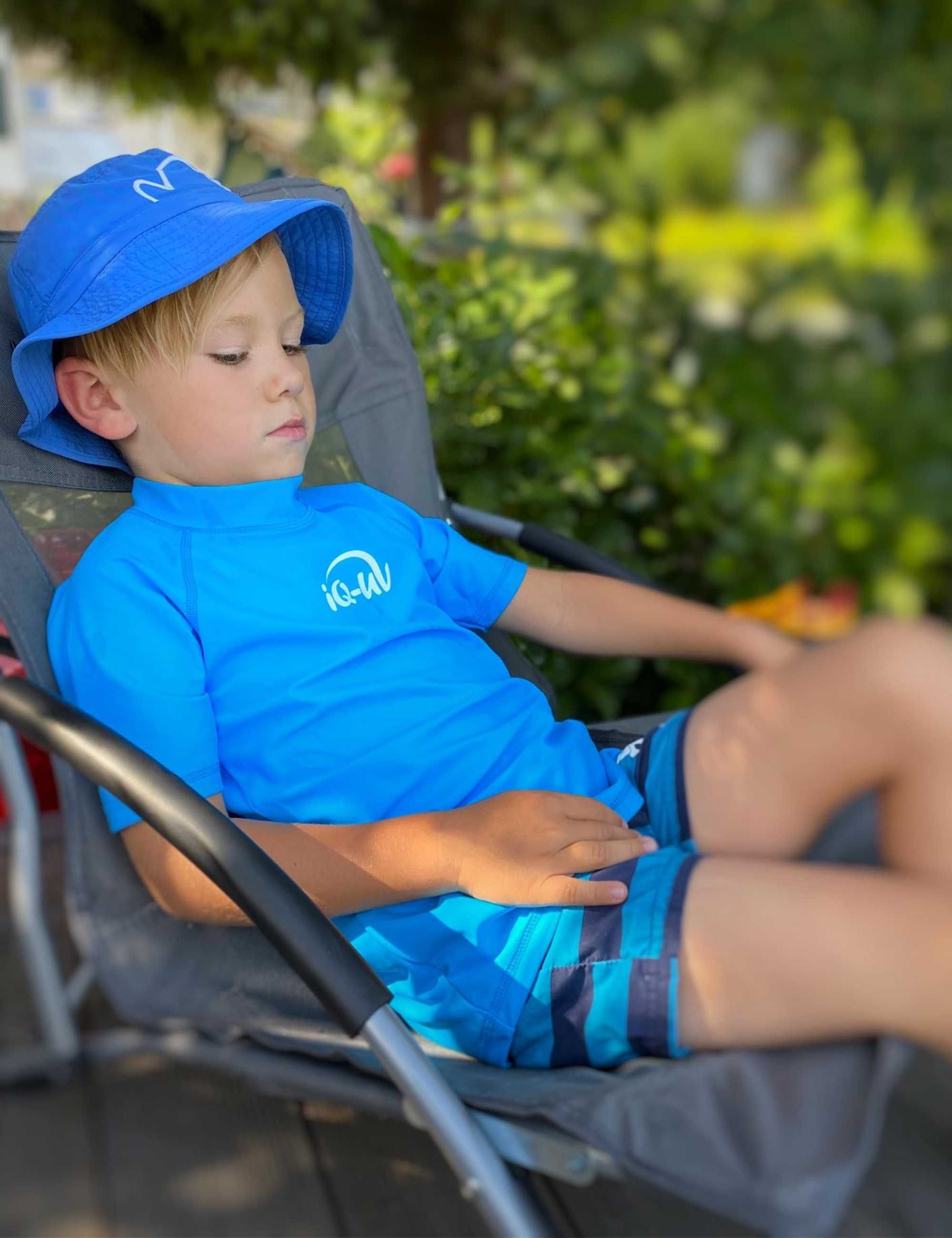 Kind in blauem Kurzarm-UV-Shirt und Hut. Weißes iQ-UV-Logo. Kind sitzt in Liegestuhl. Hintergrund: Garten.