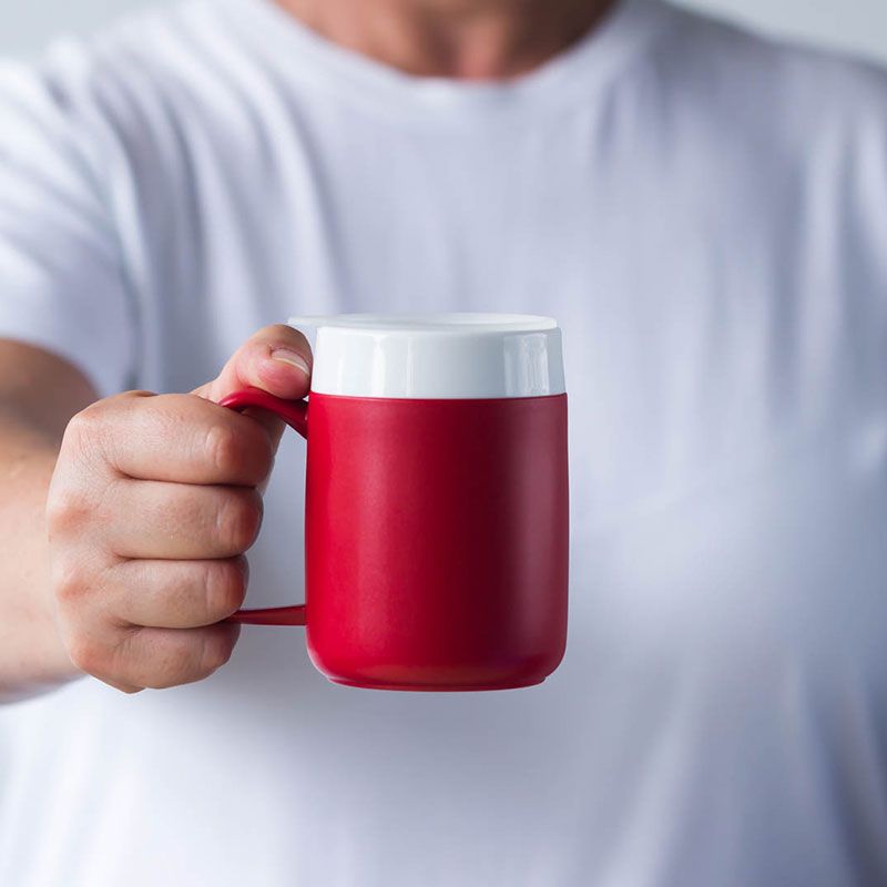 Roter Becher mit weißem Deckel und großem Henkel. Die Hand hält den Becher.