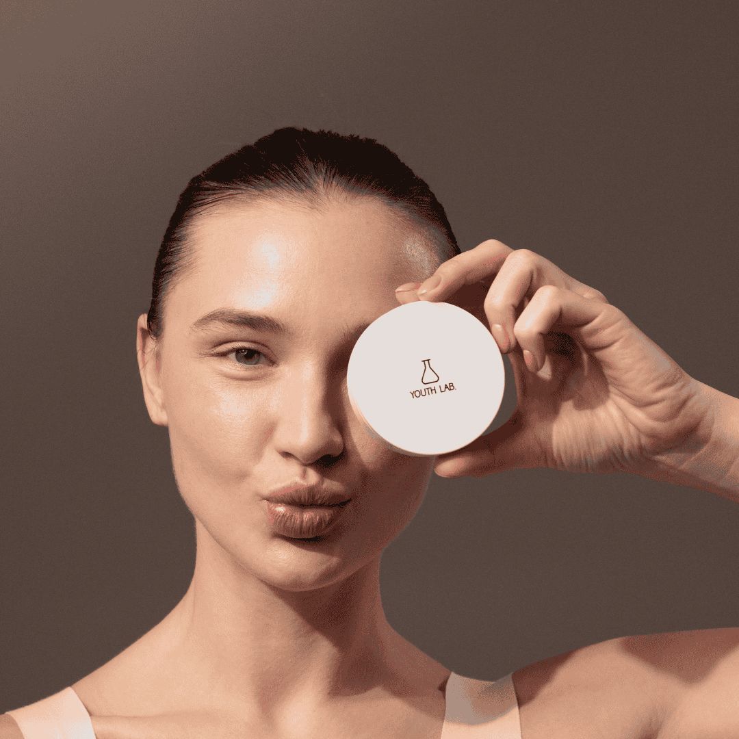 Eine Frau hält eine runde, weiße Kompaktcreme vor einem Auge. Auf dem Deckel ist ein Logo zu sehen.