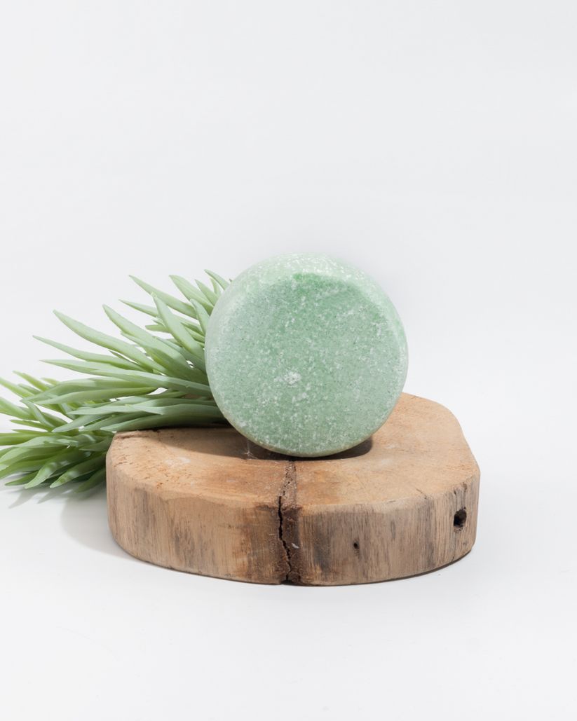 Grüner, runder Shampoo-Block auf einem Holzstück. Grünes Blattwerk daneben. Auf weißem Hintergrund.