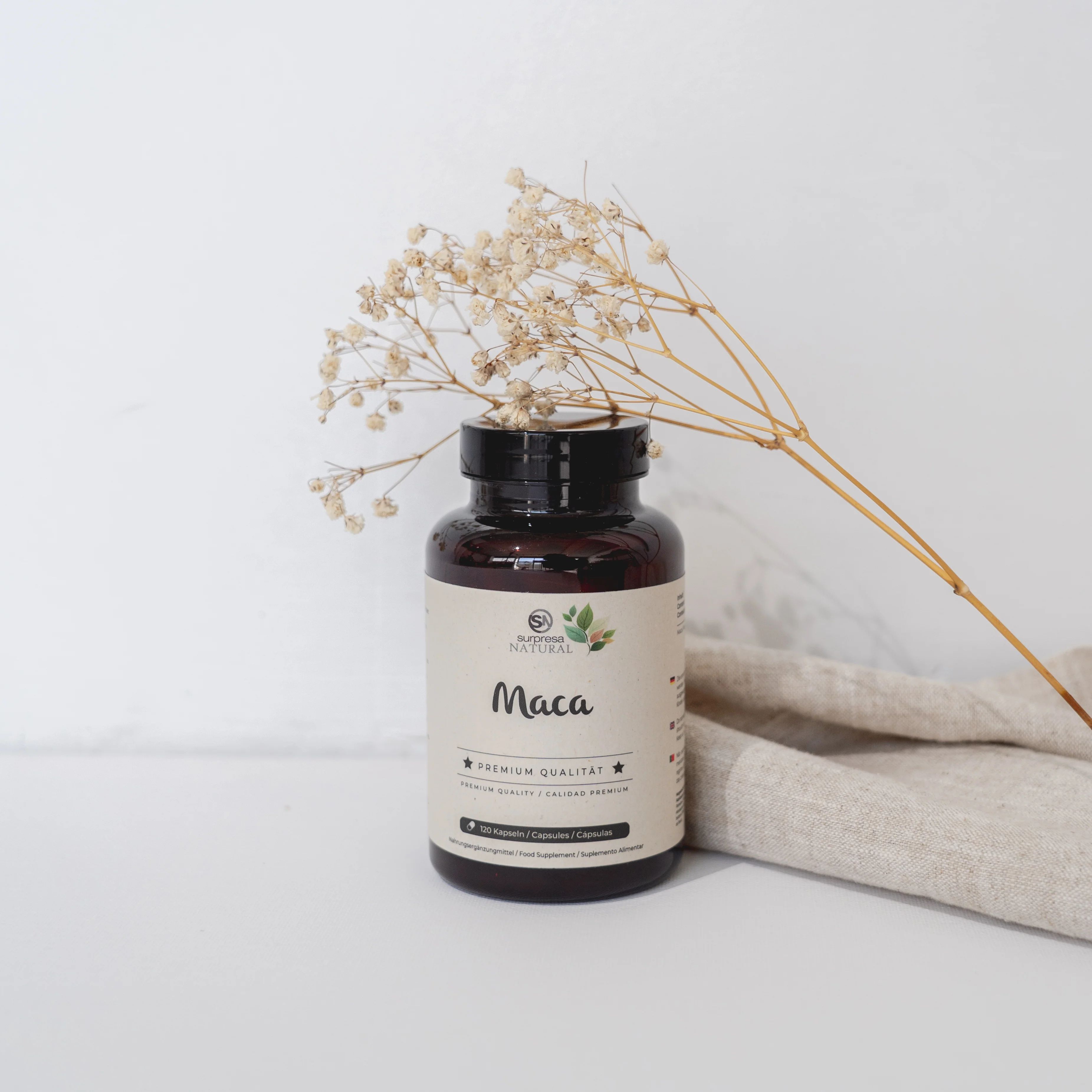 Braune Flasche mit Etikett und Aufschrift Maca. Trockenblumen und Stoff auf weißem Hintergrund.