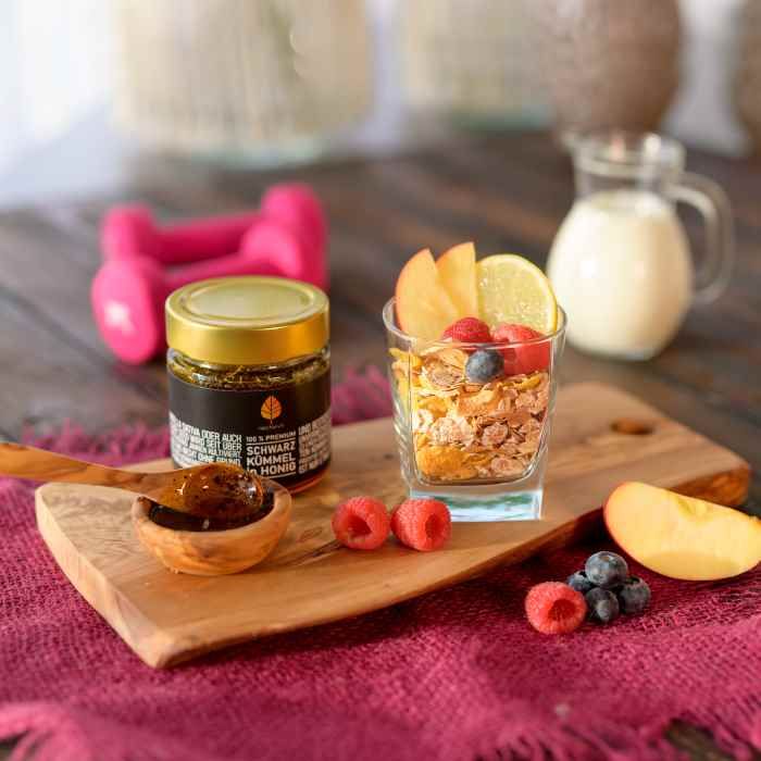 Honigglas mit goldenem Deckel neben einem Glas mit Müsli und Früchten. Löffel mit Honig und Beeren.