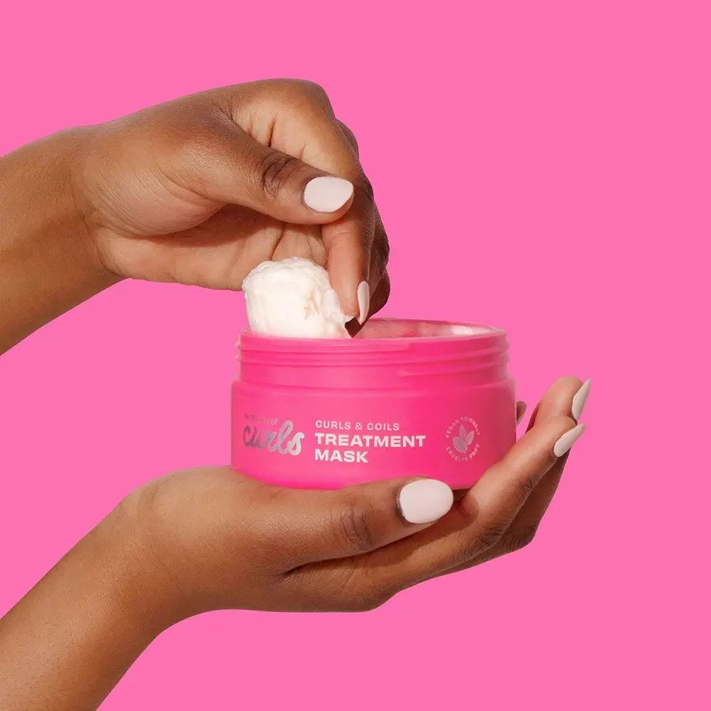 Hände halten einen rosa Tiegel mit Aufschrift 'for the love of curls', 'CURLS & COILS TREATMENT MASK'. Ein Löffel Creme wird entnommen.