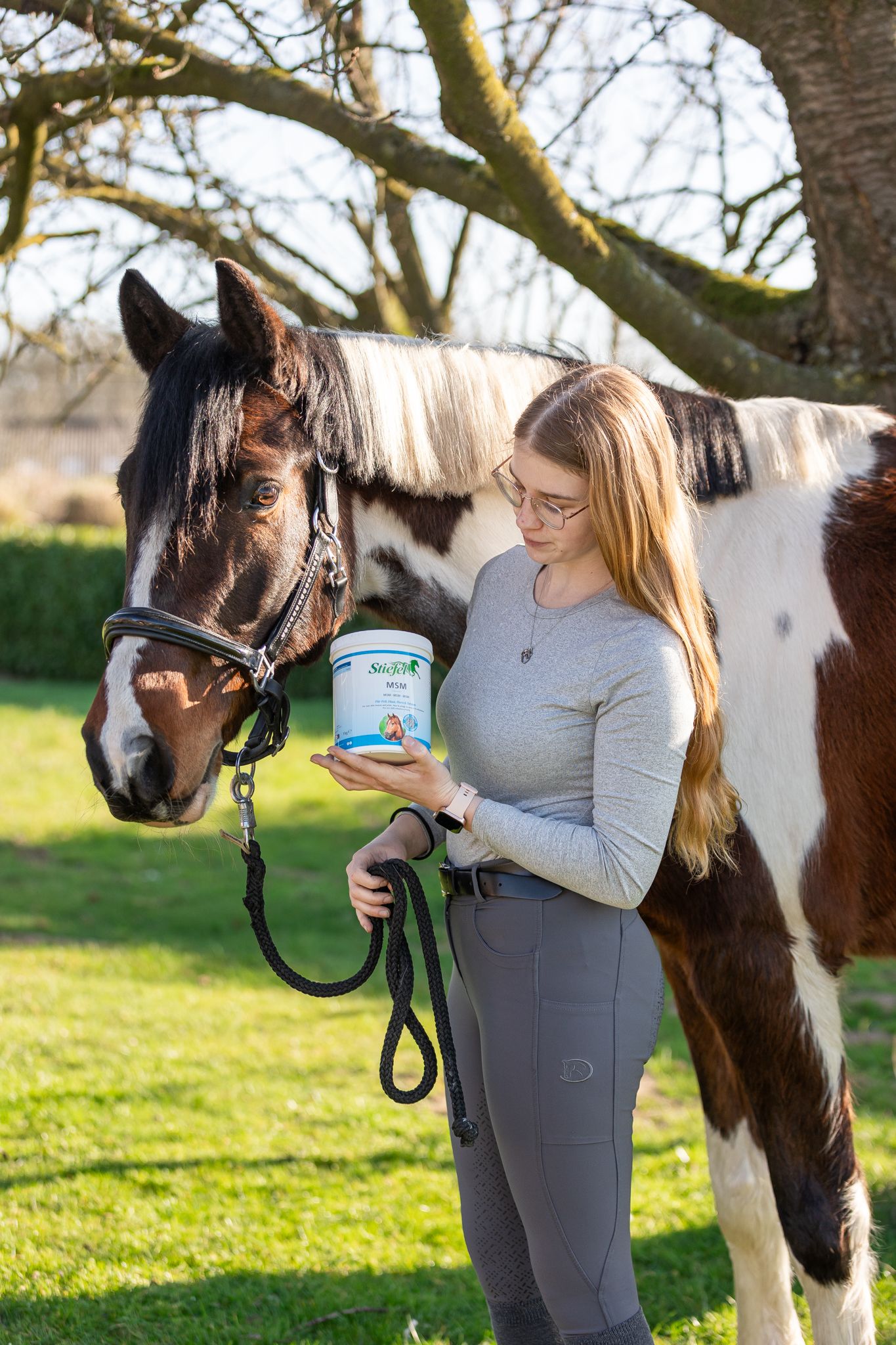 Frau hält einen Behälter vor einem Pferd. Das Produkt hat ein Etikett mit Text und einem Logo. Das Pferd trägt eine Trense.