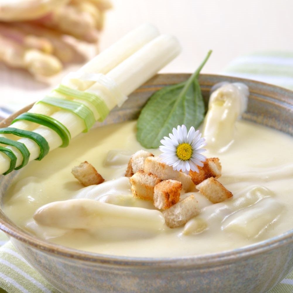 Schüssel mit Spargelsuppe, garniert mit Spargel, Croutons und einer Gänseblümchen. Grüne und weiße Dekoration.