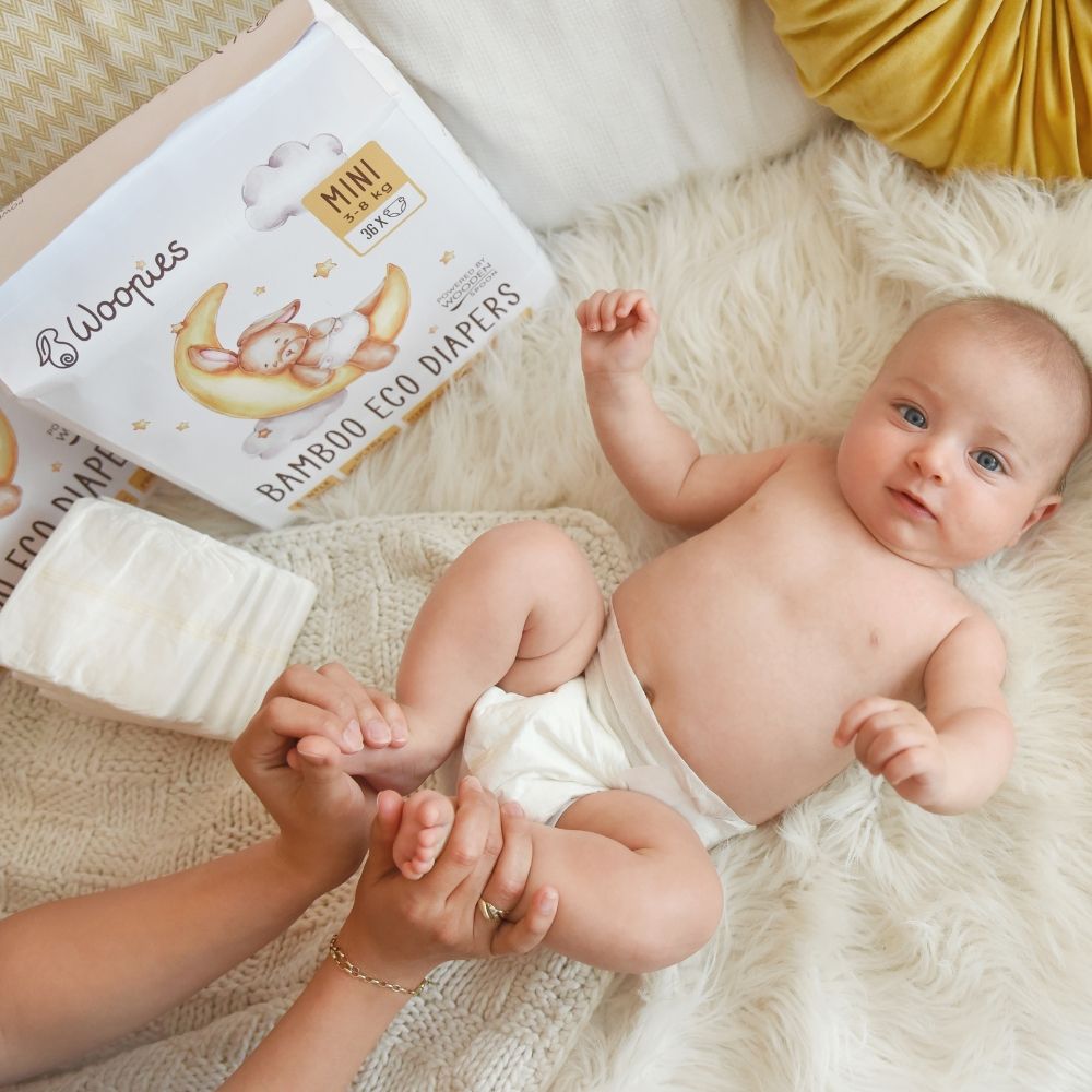 Baby liegt auf dem Rücken, Windel an. Im Hintergrund Verpackung mit Logo und Produktbezeichnung. Hände halten Babyfüße.