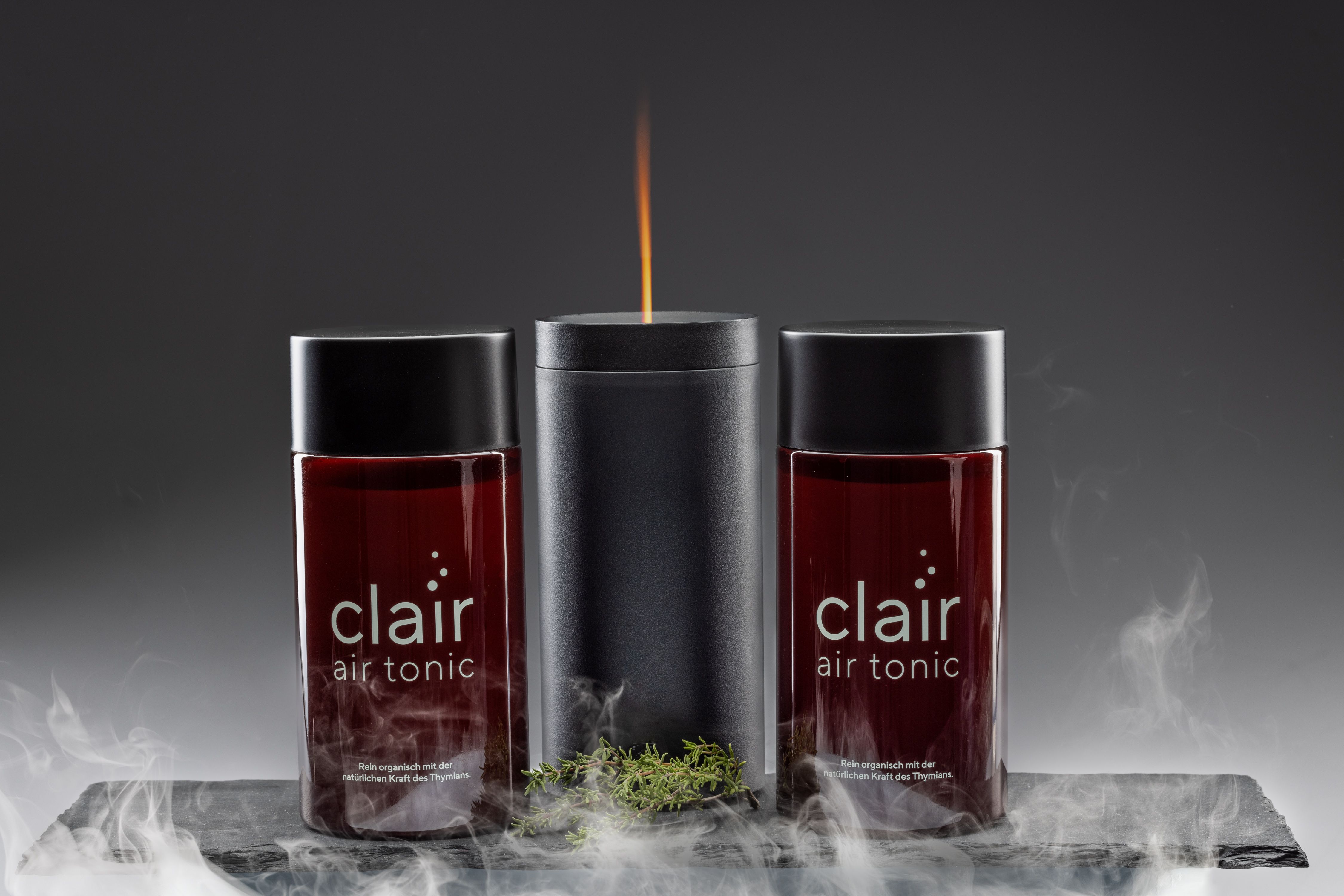 Zwei Flaschen mit schwarzem Deckel und rotem Inhalt. Aufschrift: clair air tonic. In der Mitte eine Kerze.