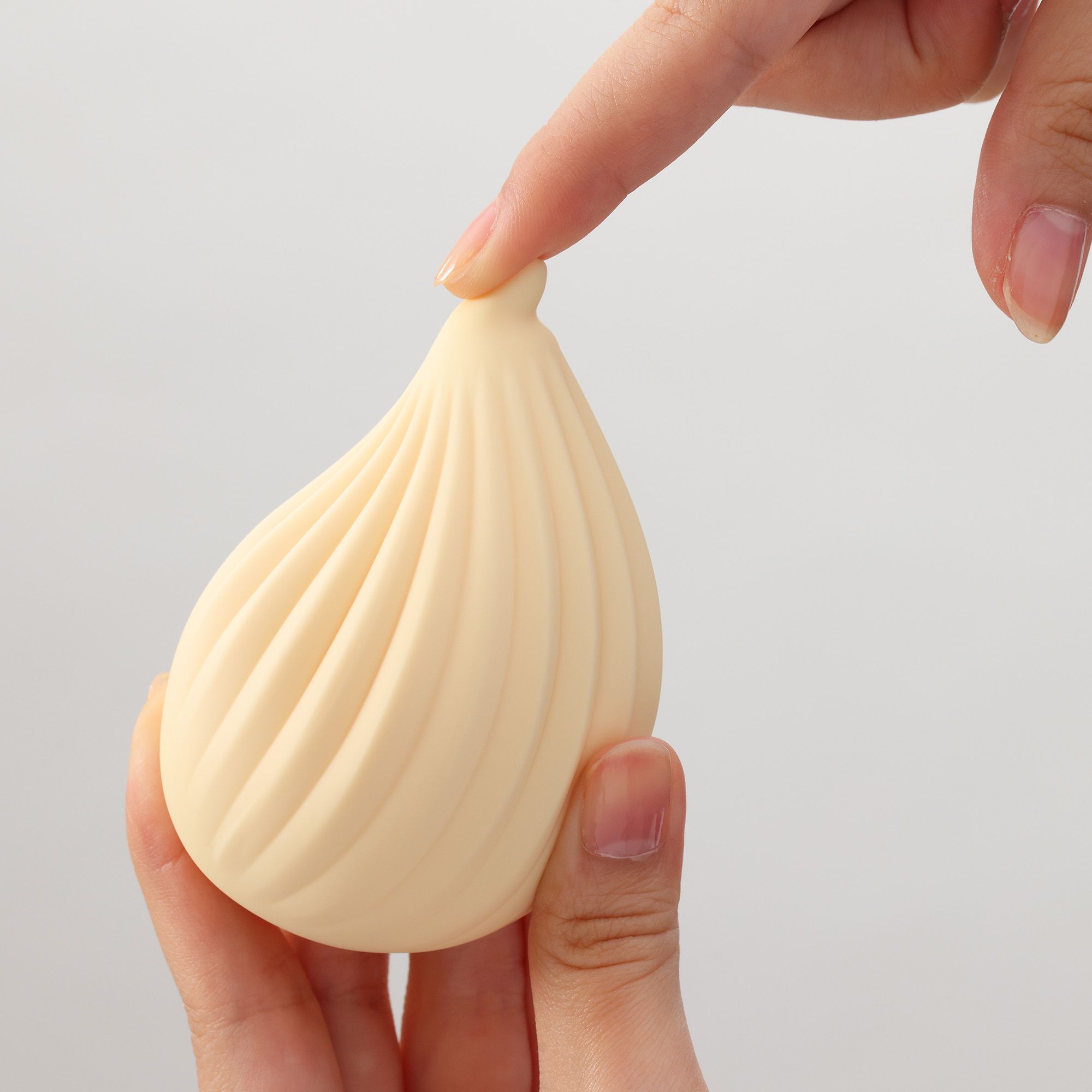 Beigefarbener Vibrator in Tropfenform. Geriffelte Oberfläche. Zwei Hände berühren das Produkt.