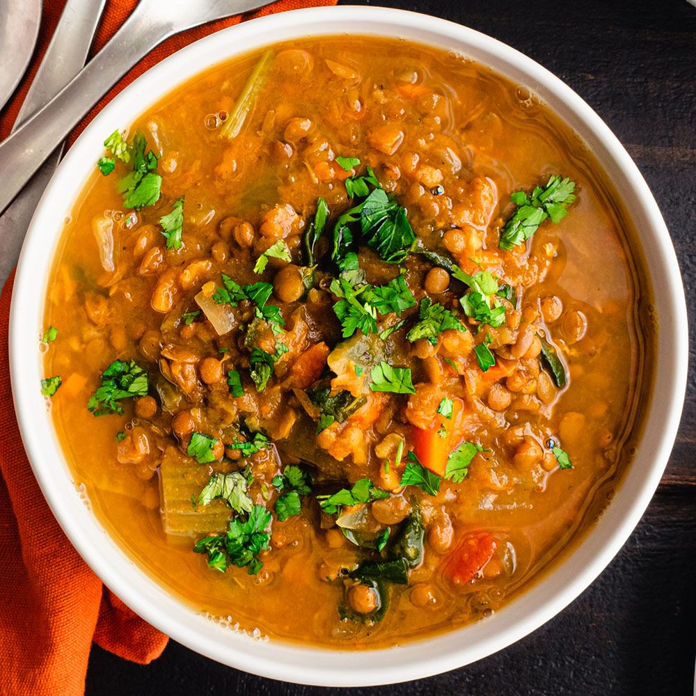 Schüssel mit Linsensuppe. Gemüse und Kräuter sichtbar. Orangefarbenes Tuch unter der Schüssel. Dunkler Hintergrund.