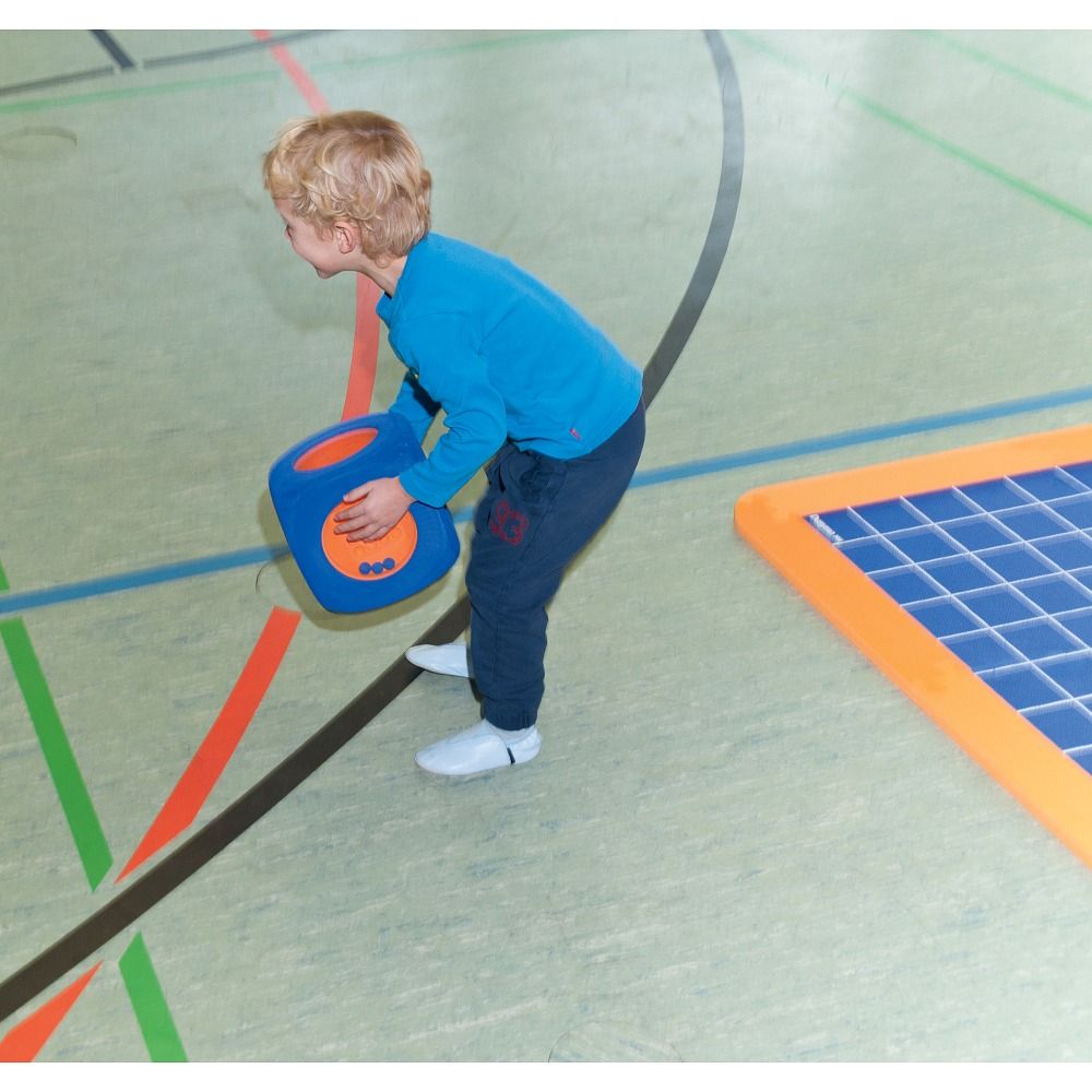 Kind hält einen blauen Würfel mit orangefarbenen Flächen. Kind steht in einer Sporthalle.