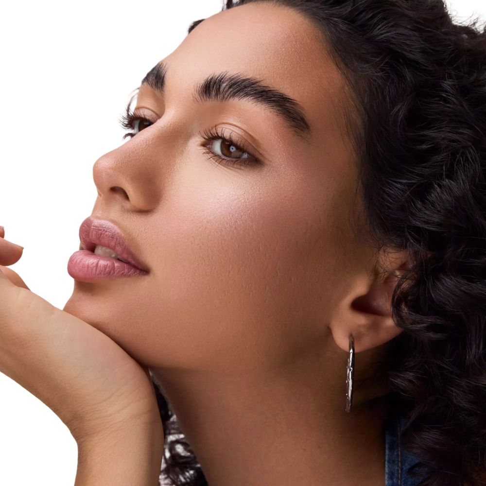 Porträt einer Frau mit gebräunter Haut. Sie trägt silberne Creolen. Ihr Gesicht ist leicht geschminkt, mit Highlighter.