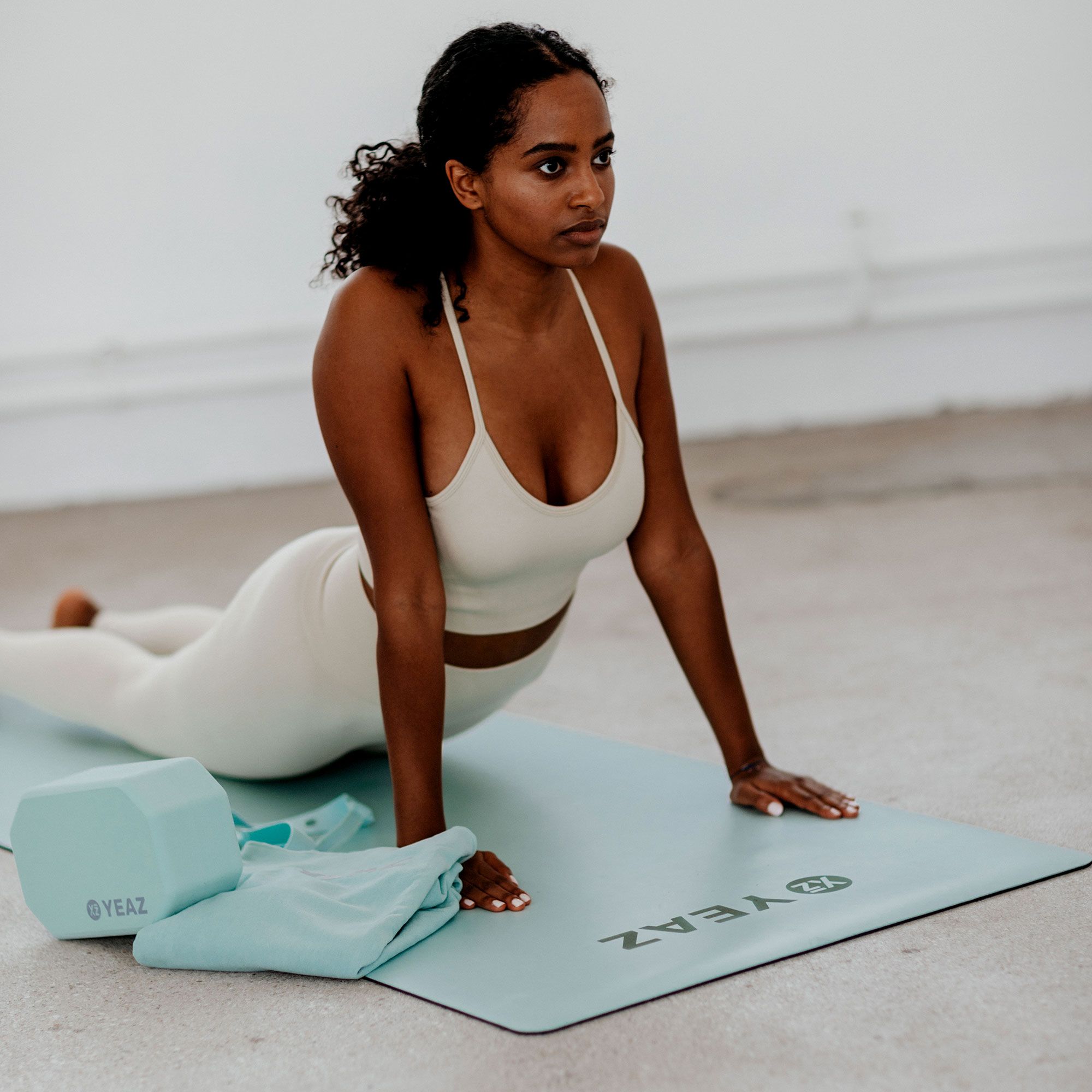 Frau macht Yoga auf hellgrüner Matte. Matte mit Logo und Schriftzug YEAZ. Yogablock und Handtuch daneben.