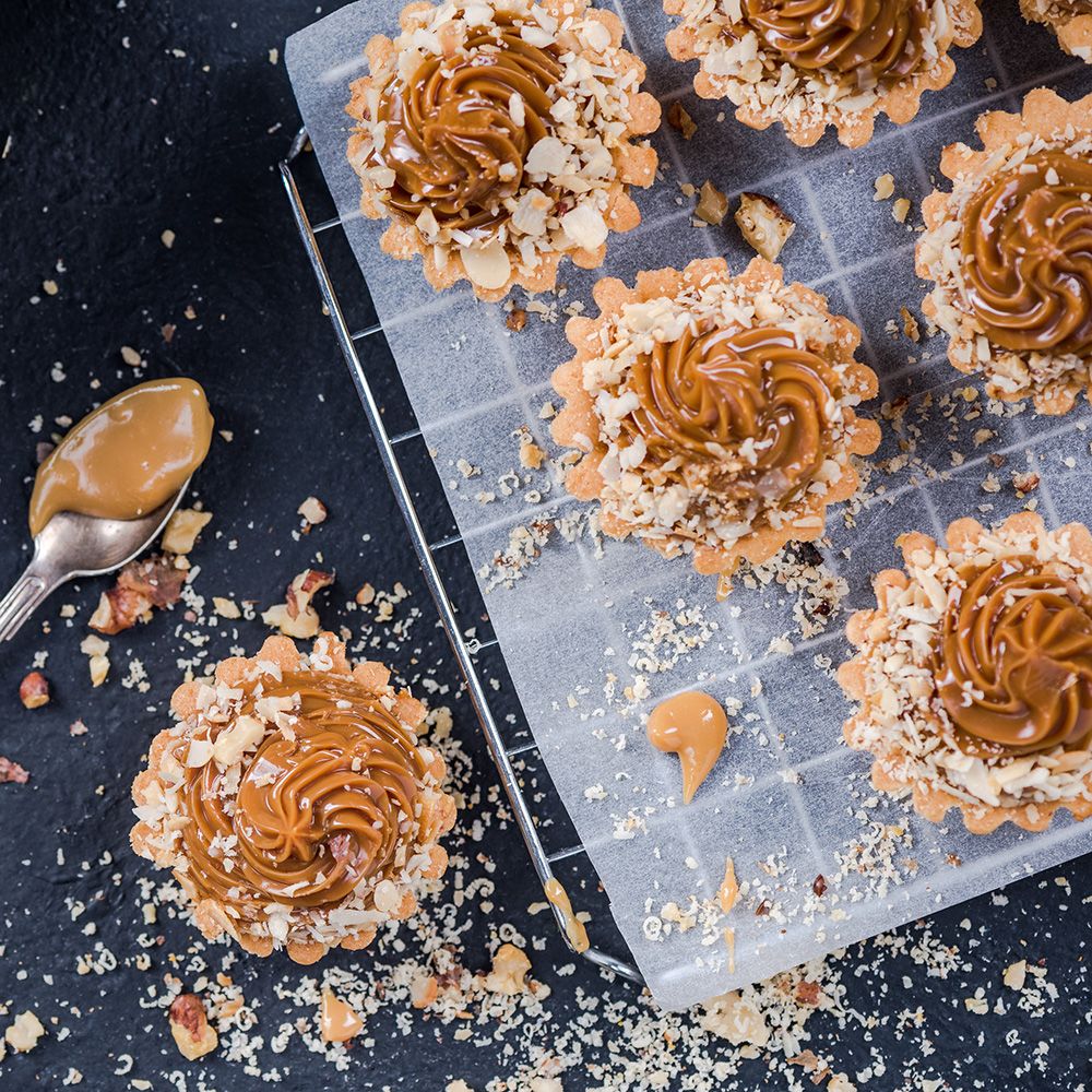 Sechs Tartes mit Karamellfüllung und Nüssen auf einem Rost. Löffel mit Karamell daneben.