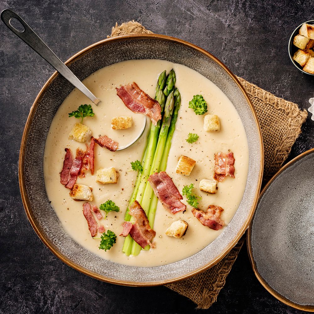 Cremige Suppe mit Spargel, Speck und Croutons in einer Schüssel. Draufsicht, Löffel in der Suppe.