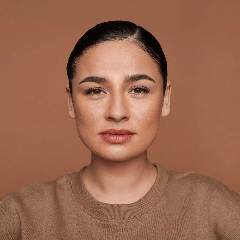 Porträt einer Frau mit gebräunter Haut. Sie trägt ein braunes Oberteil und hat dunkle Haare. Ihr Gesicht ist ebenmäßig geschminkt.