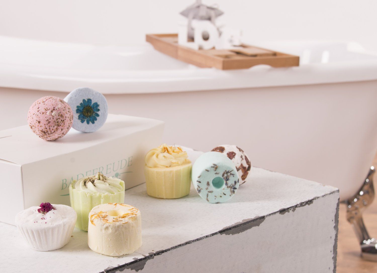 Badebomben und -kuchen auf einer weißen Oberfläche, im Hintergrund eine Badewanne. Aufdruck "Badefreude" auf der Box.