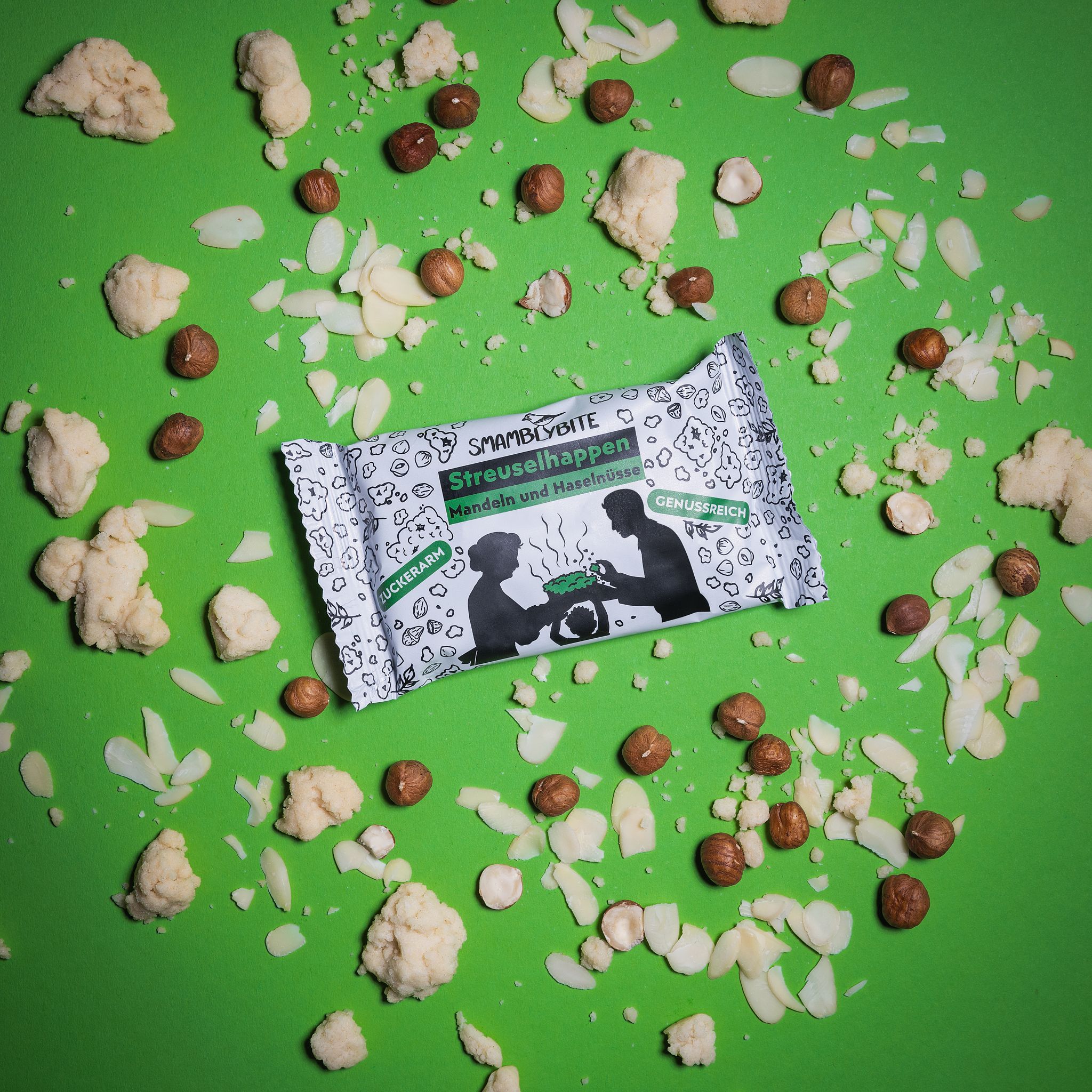 Streuselhappen-Packung auf grünem Hintergrund. Haselnüsse, Mandeln und Streusel verstreut. Aufdruck: SMAMBLYBITE, Streuselhappen.
