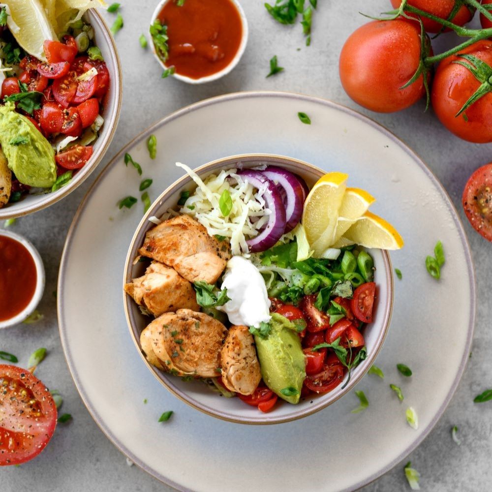 Schüssel mit Hähnchen, Tomaten, Avocado, Zwiebeln, Zitrone und Kräutern. Auf hellem Teller. Zwei kleine Schalen mit Soße. Draufsicht.