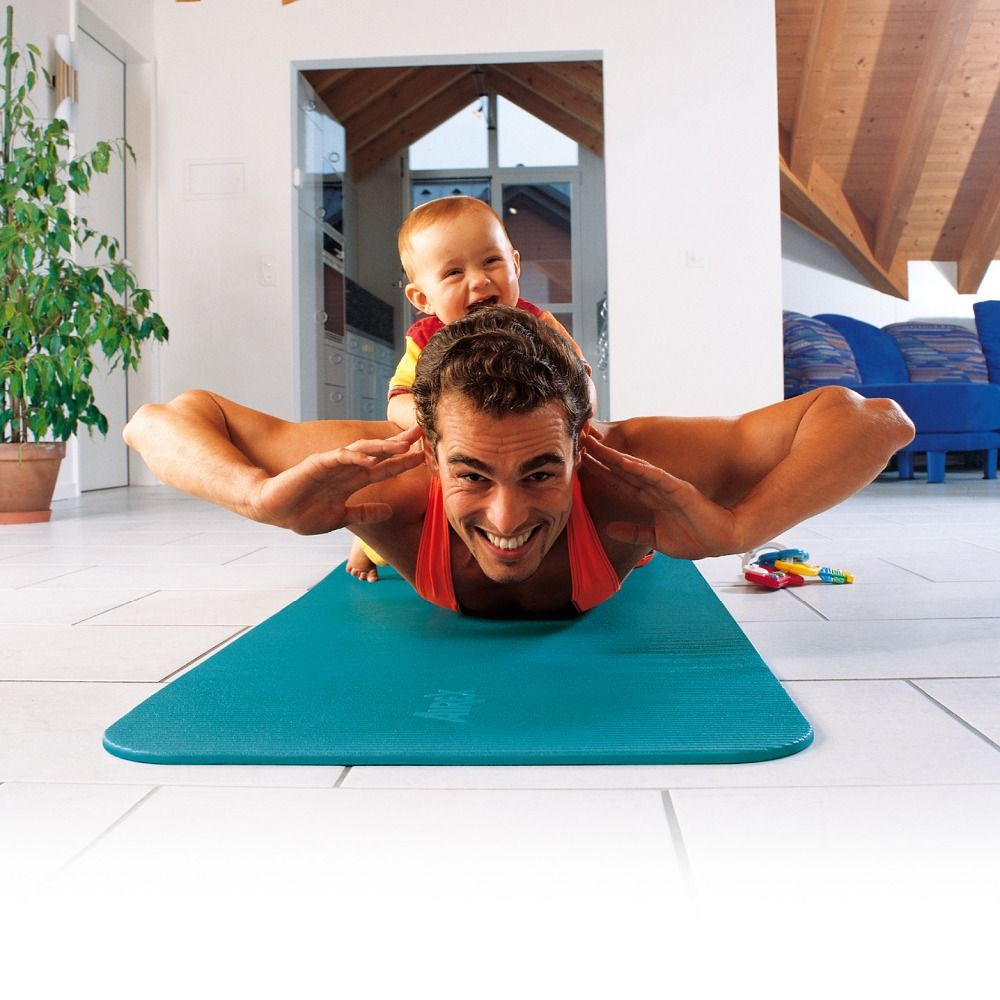 Türkisfarbene Gymnastikmatte. Mann liegt auf der Matte, Baby sitzt auf seinem Rücken. Matte im häuslichen Umfeld.