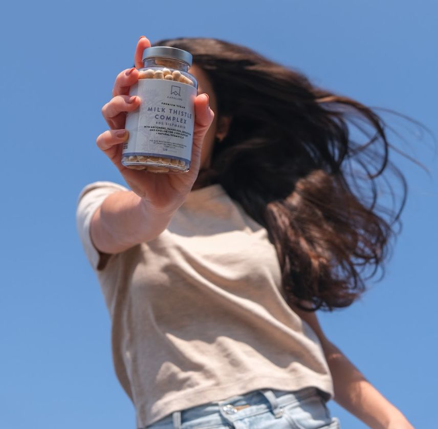 Person hält eine Kapsel-Glasflasche. Aufschrift: Milk Thistle Complex. Blauer Himmel.