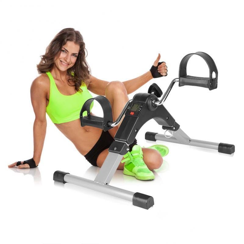 Mini-Heimtrainer. Frau sitzt daneben. Schwarz-silberfarbenes Gerät mit Pedalen und digitalem Display. Für Arme und Beine.