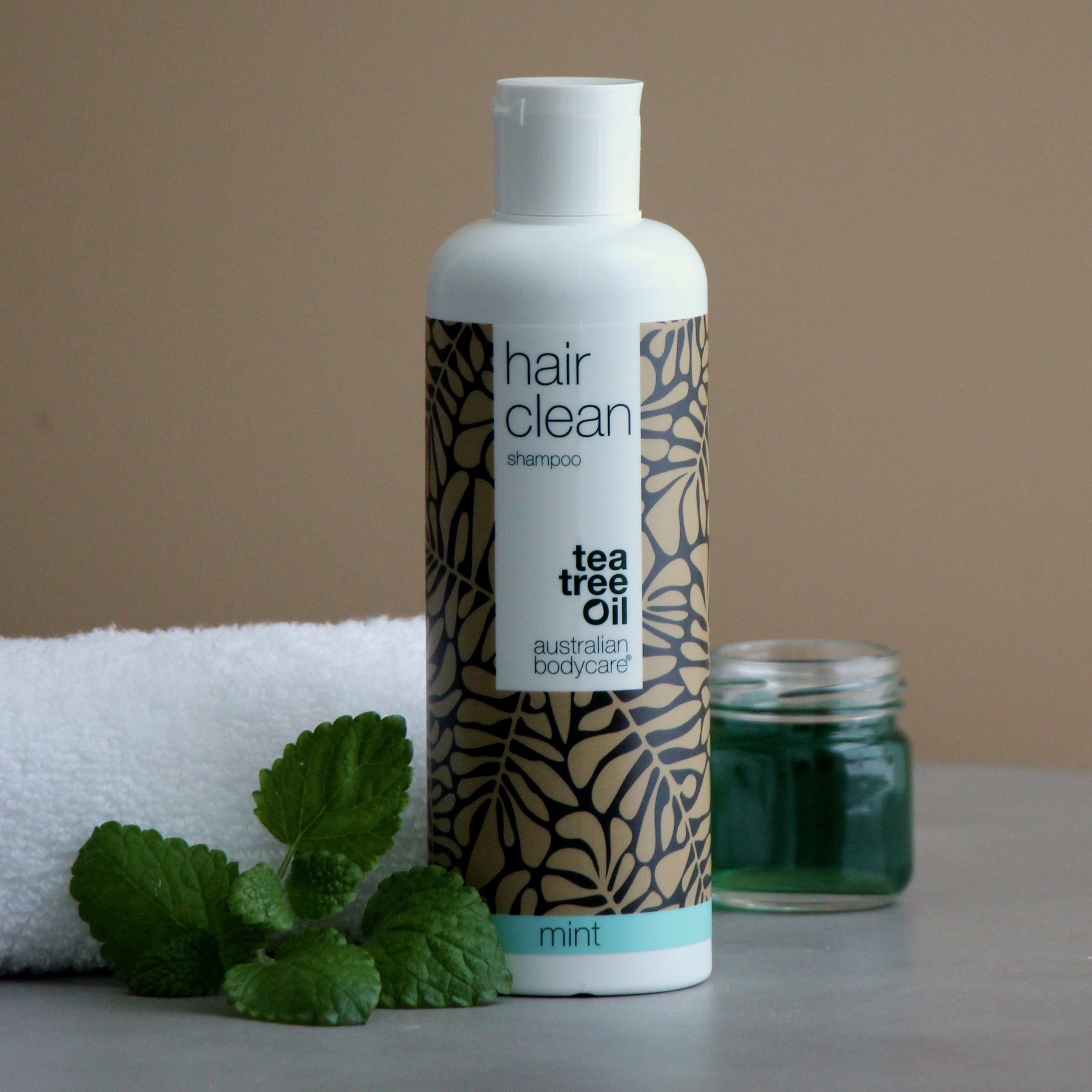 Shampoo-Flasche mit floralem Muster, Schriftzug 'hair clean shampoo', 'tea tree oil' und 'mint'. Daneben ein Handtuch, Minze und ein Glas.