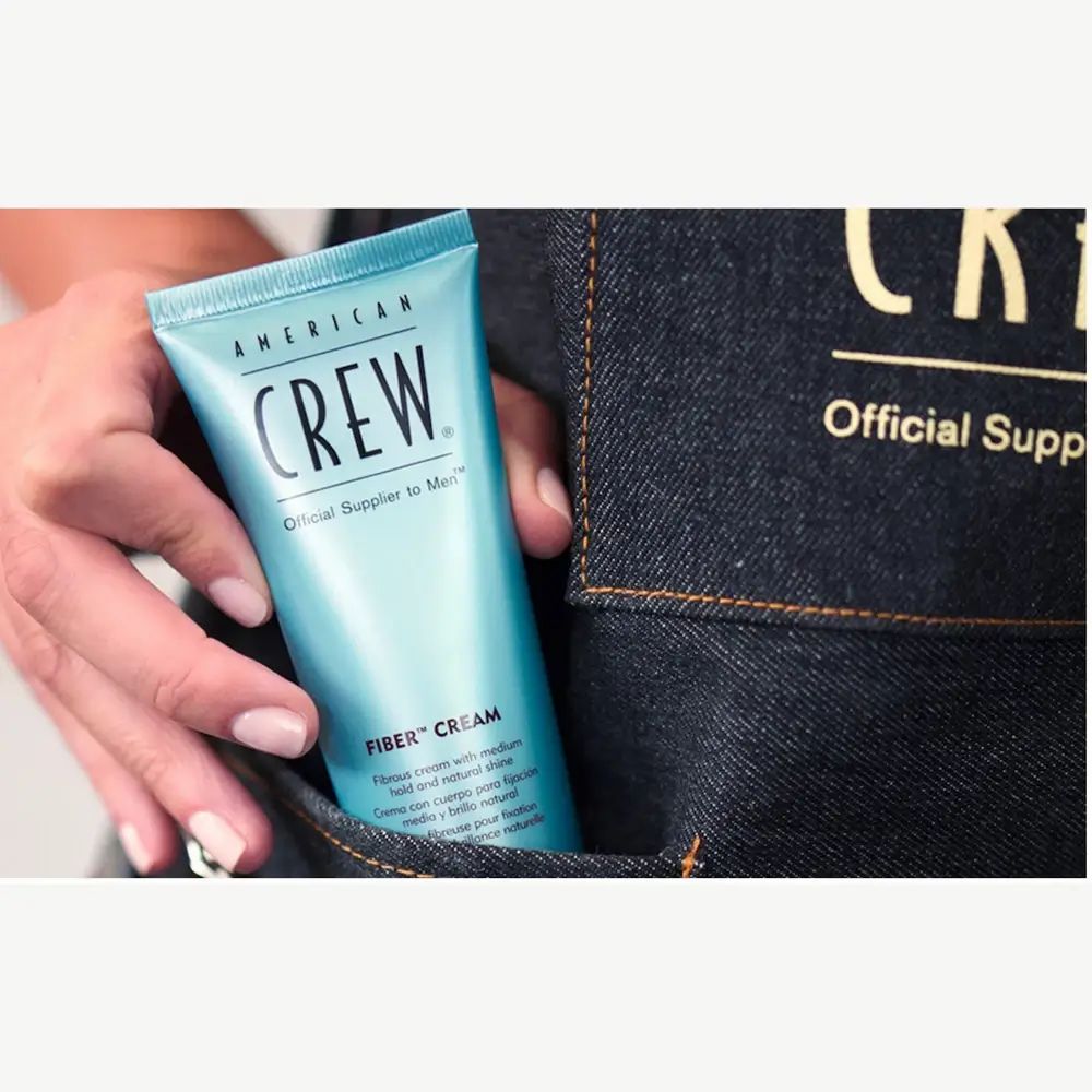 Türkisfarbene Tube mit schwarzem Deckel. Aufschrift: American Crew, Fiber Cream. In Hand, vor Jeans-Tasche.