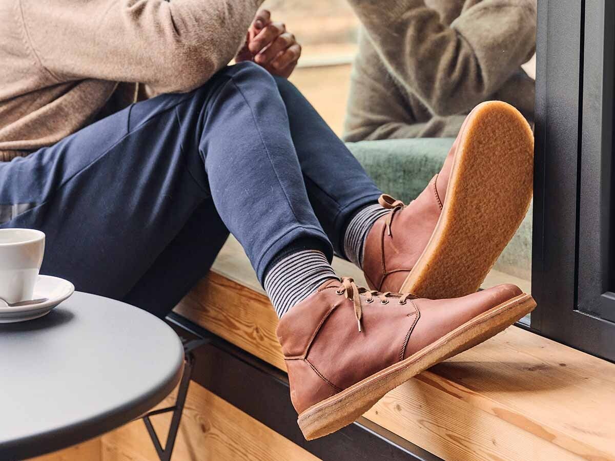 Person sitzt mit braunen Leder-Stiefeletten auf einer Fensterbank. Blaue Hose, gestreifte Socken, Kaffeetasse.